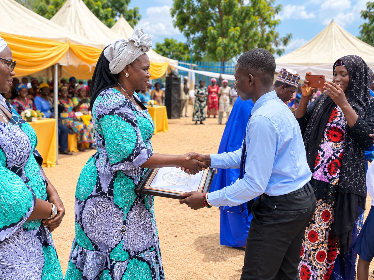 The Proprietress of Greater Future Academy presenting a framed award and shaking hands with a pupil at an outdoor ceremony